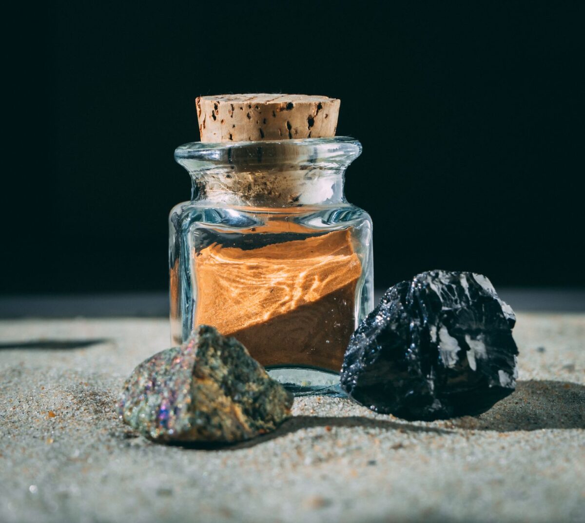 shilajit for stamina – A glass jar with cork lid next to two stones on a sandy surface, under sunlight.