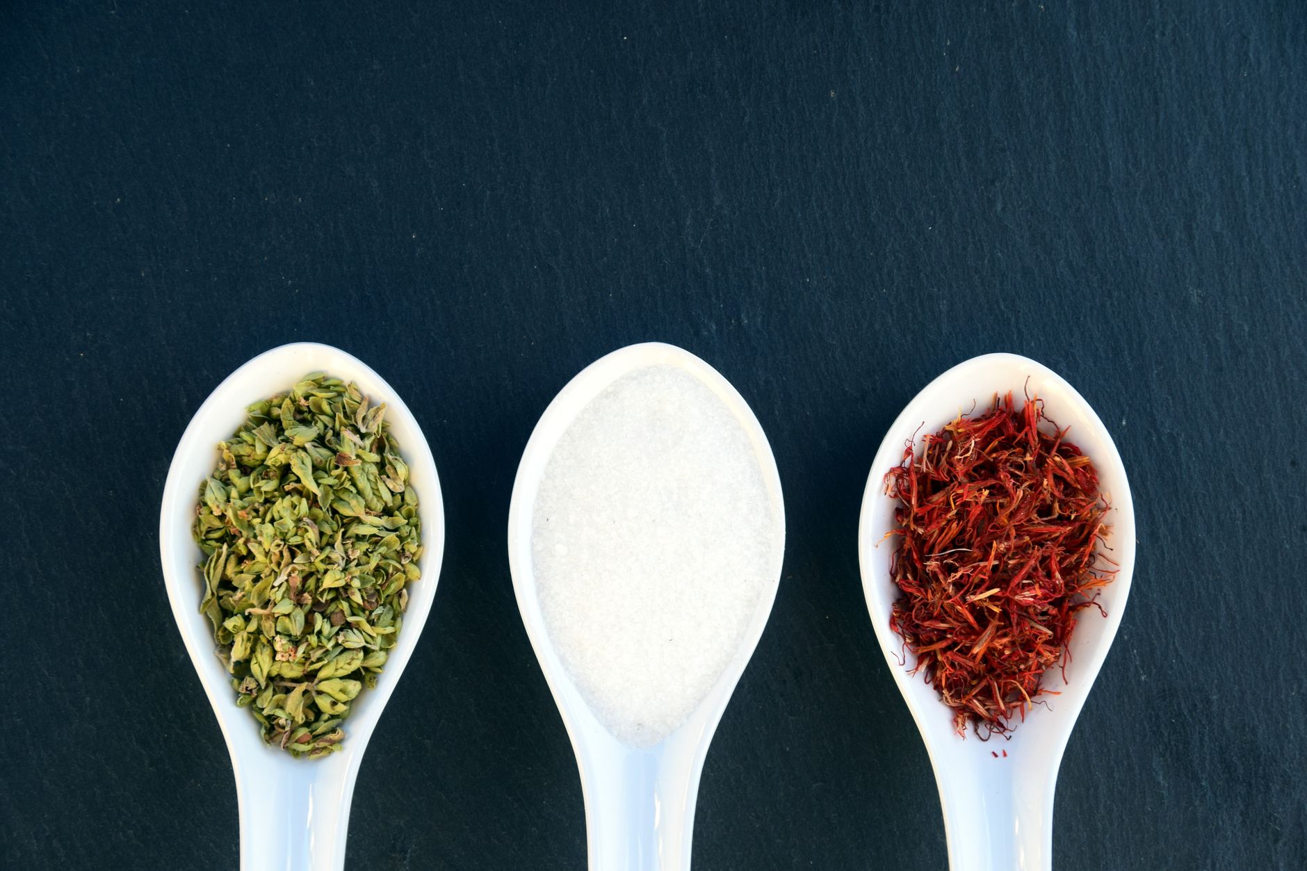 زعفران ریٹ پاکستان – Three ceramic spoons with herbs, salt, and saffron on dark slate background.